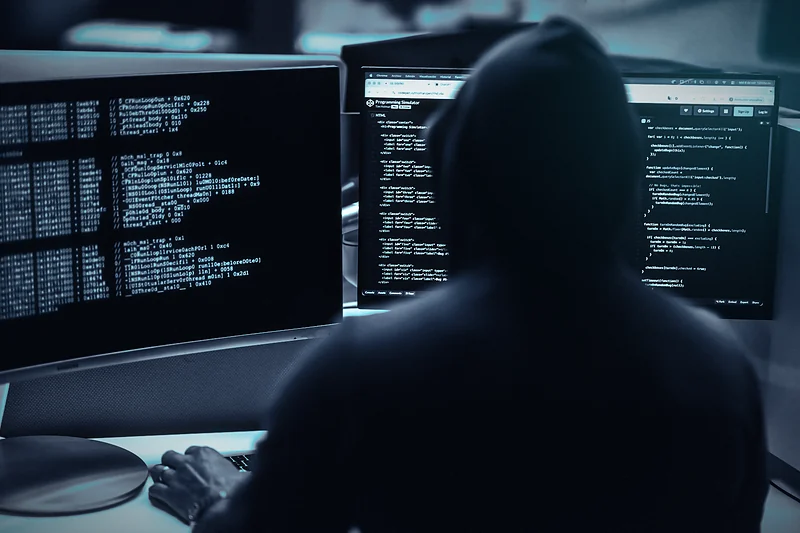 A hooded hacker seen from behind, typing at a desk in front of screens full of code